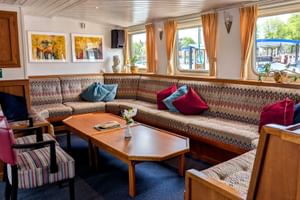 Gemütliche Schiffslounge mit Eckbank, bunten Kissen, Holzcouchtischen und großen Fenstern mit orangefarbenen Vorhängen mit Blick auf einen Kanal.