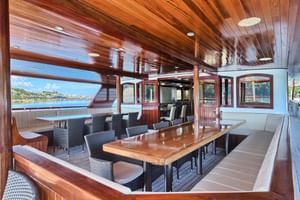 Innenbereich des Außendecks der Andela Lora mit Holzdecke, Esstischen, Korbstühlen und großen Fenstern mit Küstenblick.