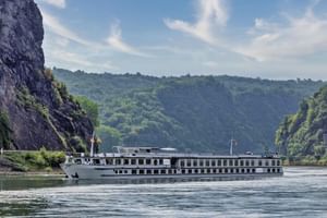 MS Veloria river cruise ship sailing on calm water between steep rocky cliffs covered with green vegetation under a blue sky.