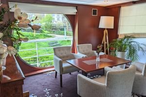 Elegante Bibliothek-Lounge auf der SE Manon mit beigen Sesseln um einen Holztisch, großes Panoramafenster mit Blick auf grüne Uferland.