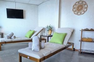 Entspannungsraum mit zwei Holzliegen mit weißen Kissen und grünen Polstern, Wandfernseher, dekorativer Wandkunst und Regal.