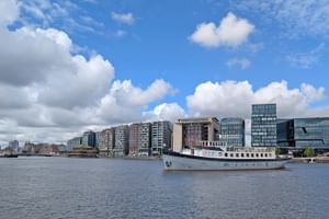 Weißes Passagierschiff Floor van Aemstel auf ruhigem Wasser mit modernen Gebäuden und blauem Himmel mit weißen Wolken im Hintergrund.