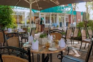 Außenterrasse im Upstalsboom Parkhotel Emden mit gedeckten Tischen, Korbstühlen unter Sonnenschirmen, umgeben von Grün und Hotelgebäude.