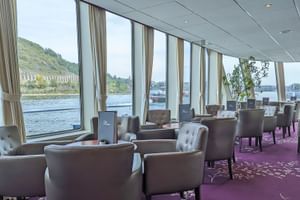 Moderne Lounge auf der SE Manon mit grauen Sesseln, lila Teppich und bodentiefen Fenstern mit Blick auf einen Fluss und grünen Hang.