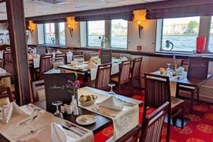 Innenraum des MS Normandie Schiffsrestaurants mit gedeckten Tischen, dunklen Holzstühlen, rotem Teppich und großen Fenstern mit Wasserblick.
