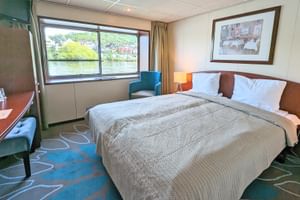 Helle Kabine auf der SE Manon mit Doppelbett, Fenster mit Flussblick, Schreibtisch, Sessel und Nachttischlampen. Grüner Teppich und weiße Wände.