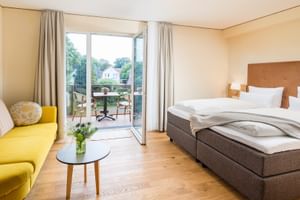 Helles Hotelzimmer im Parkhotel Jever mit Doppelbett, gelbem Sofa, Holzboden und Balkontür zu einem Sitzbereich im Freien.