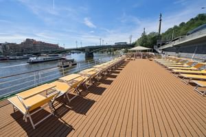 Holz-Sonnendeck der MS Florentina mit gelben gepolsterten Liegen in Reihen. Flussblick mit Brücke und historischen Gebäuden unter blauem Himmel.