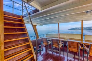 Außenbereich des Schiffes Linda mit Holztreppe, Esstisch, Stühlen und Panorama-Meerblick durch große Fenster.