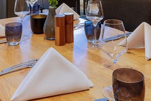 Elegant gedeckte Restauranttische mit weißen Servietten, Weingläsern, Wassergläsern und kupferfarbenen Accessoires auf hellem Holz.