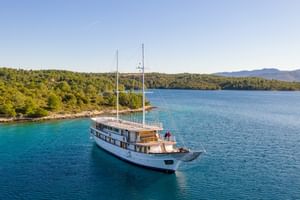 Luftaufnahme der weißen Segelyacht Magellan vor Anker im türkisfarbenen Wasser einer bewaldeten kroatischen Bucht mit Bergen im Hintergrund.