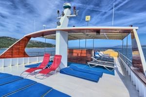 Sun deck of MY Andela Lora with blue and red loungers under a wooden canopy. Croatian coastline visible in the background.