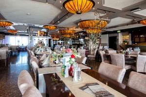 Innenraum des Hotel Old Dutch Restaurants in Volendam mit beigen gepolsterten Stühlen, gedeckten Tischen mit Blumen und orangefarbenen Pendelleuchten.