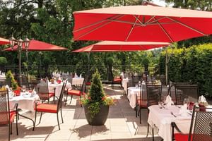 Außenterrasse des Victors Residenz Hotels mit weiß gedeckten Tischen, Korbstühlen, roten Sonnenschirmen und Blumenkübeln umgeben von Bäumen.