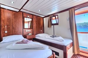 2-Bett-Kabine an Bord der Andela Lora mit zwei Einzelbetten, Holzvertäfelung, Bullauge mit Meerblick und beigen Vorhängen.