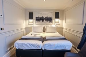 Elegant junior suite on MS MAGNIFIQUE IV with twin beds, white linens, decorative wall panels, and a framed cityscape artwork above the headboard.