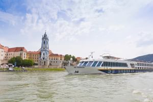 Weißes Flusskreuzfahrtschiff SE Manon auf der Donau mit dem blauen barocken Kirchturm von Dürnstein und historischen Gebäuden am Ufer.