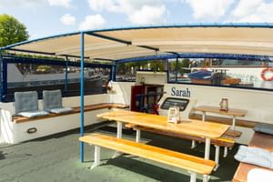 Sun deck of MS Sarah with wooden picnic tables, bench seating, and blue canopy awning. The ship's wheelhouse is visible in the background.