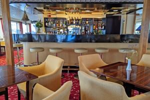 Innenraum des MS Normandie Salons mit Bar, Flaschen, Barhockern, cremefarbenen Sesseln um dunkle Holztische und rotem gemustertem Teppich.