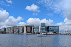 Weißes Passagierschiff Floor van Aemstel am modernen Ufer mit zeitgenössischen Glas- und Betongebäuden unter blauem bewölktem Himmel.