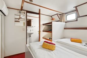 Kompakte Schiffskabine mit zwei Betten, weißen Wänden mit Holzverkleidung, Bullauge, Waschbecken, Klimaanlage und bunten Handtüchern.