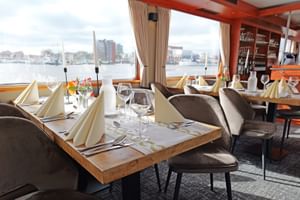 Elegantes Restaurant an Bord der MS Fluvius mit gedeckten Tischen, grauen Polsterstühlen und großen Fenstern mit Blick auf einen Hafen.