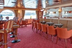 Innenraum des MS Switzerland Salons und Bar mit orangefarbenen Stühlen, Barhockern, runden Tischen und großen Bogenfenstern mit Blick aufs Wasser.
