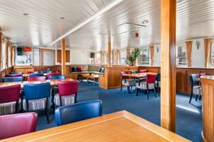 Geräumiges Restaurant an Bord der Floor van Aemstel mit blauem Teppich, Holzverkleidung, bunten Stühlen in Rot und Blau und weißer Decke mit Fenstern.