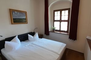 Helles Hotelzimmer mit Doppelbett, weißer Bettwäsche, Fensternische mit dunklen Vorhängen, Landschaftsgemälde und Holzboden.