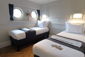 Twin cabin on MS MAGNIFIQUE IV with two single beds, white linens, round portholes with blue curtains, and wooden flooring.