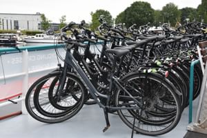 Multiple black bicycles parked in bike racks on the white deck of MS Flora ship, with boats and harbor buildings visible in background.