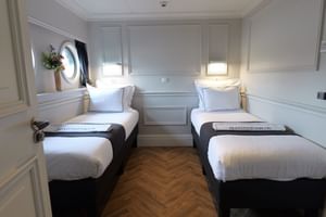 Twin cabin on MS MAGNIFIQUE IV with two single beds, white linens, porthole window, built-in storage, and parquet flooring.