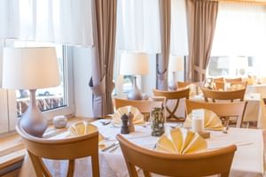Eleganter Speisesaal im Hotel Seeblick mit runden Tischen mit weißen Tischdecken, gelben Servietten und Holzstühlen nahe großen Fenstern.