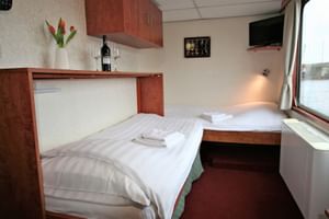 Doppelkabine auf der MS Fluvius mit zwei Betten, weißer Bettwäsche, Holzmöbeln, großem Fenster mit Flussblick und kleinem Wandfernseher.