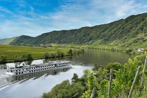 MS Olympia Flusskreuzfahrtschiff auf ruhigem Fluss mit grünen Weinberghängen, bewaldeten Hügeln und kleinem Dorf im Hintergrund unter blauem Himmel.