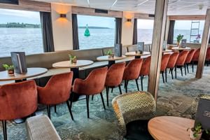 Moderne Lounge auf der MS Princess mit orangefarbenen Samtstühlen, runden Tischen und Panoramafenstern mit Blick auf einen Fluss.
