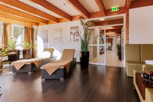 Spa-Ruheraum mit Holzbalken, beigen Liegen, dunklem Boden und gläsernem Saunaeingang. Dekoriert mit Pflanzen und Tageslicht.