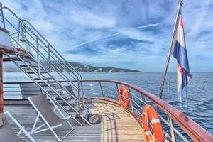 Stern deck of MY Andela Lora with wooden flooring, metal stairs, orange life rings, and Croatian flag. Coastal hills visible across calm sea.