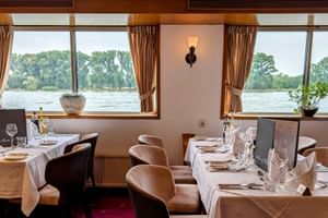 Elegantes Restaurant an Bord der MS Olympia mit weiß gedeckten Tischen, braunen Lederstühlen und großen Fenstern mit Blick auf einen Fluss.