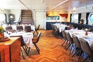 Elegantes Restaurant-Interieur auf der MS Magnifique Vier mit Fischgrät-Holzboden, weiß gedeckten Tischen, braunen Stühlen und Bullaugen.