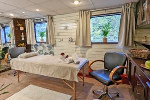 Massagesalon auf MS Prinzessin Katharina mit Massageliege, Bürostuhl, Holzschränken und Fenstern mit grünen Pflanzen auf den Fensterbänken.