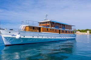 Weiße Motoryacht Linda mit Holzaufbau und blauen Akzenten vor Anker in kristallklarem blauen Wasser unter teilweise bewölktem Himmel.