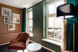 Modernes Hotelzimmer mit grünen Wänden, braunem Sessel, Stehlampe und Fensterblick. Gerahmte maritime Kunstwerke an brauner Wand.