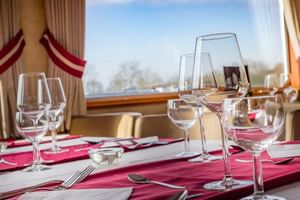 Eleganter Restauranttisch auf der MS Fluvius mit Weingläsern, Besteck und burgunderroten Servietten. Große Fenster zeigen Flussblick.