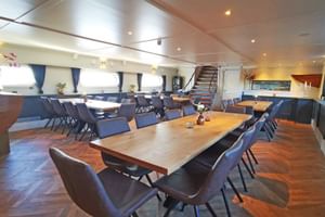 Geräumiges Restaurant an Bord der MS Magnifique Vier mit Holztischen, blau gepolsterten Stühlen, Fenstern mit Vorhängen und einer Treppe im Hintergrund.