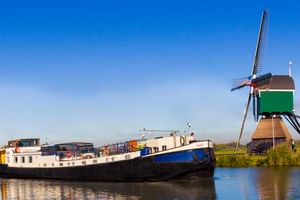 Blue and white cargo barge MS Sarah moored on a canal beside a traditional Dutch windmill with green body and brown base under clear sky.