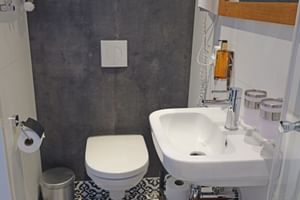 Kompaktes Badezimmer auf der MS Magnifique Vier mit weißer Toilette und Waschbecken, dunkelgrauen Wandfliesen, gemusterten Bodenfliesen.