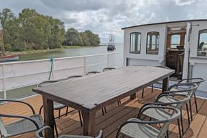 Holz-Sonnendeck mit Esstisch und Stühlen auf Schiff Floor van Aemstel. Fluss und Bäume im Hintergrund unter bewölktem Himmel.