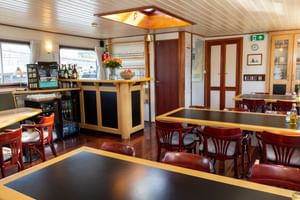 Interior of MS Sarah restaurant showing dining tables with dark tops, red wooden chairs, a central bar area, and windows along the side.