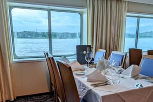 Eleganter Restauranttisch auf der SE Manon mit weißer Tischdecke, Weingläsern und Servietten. Große Fenster zeigen Flussblick mit Ufer.